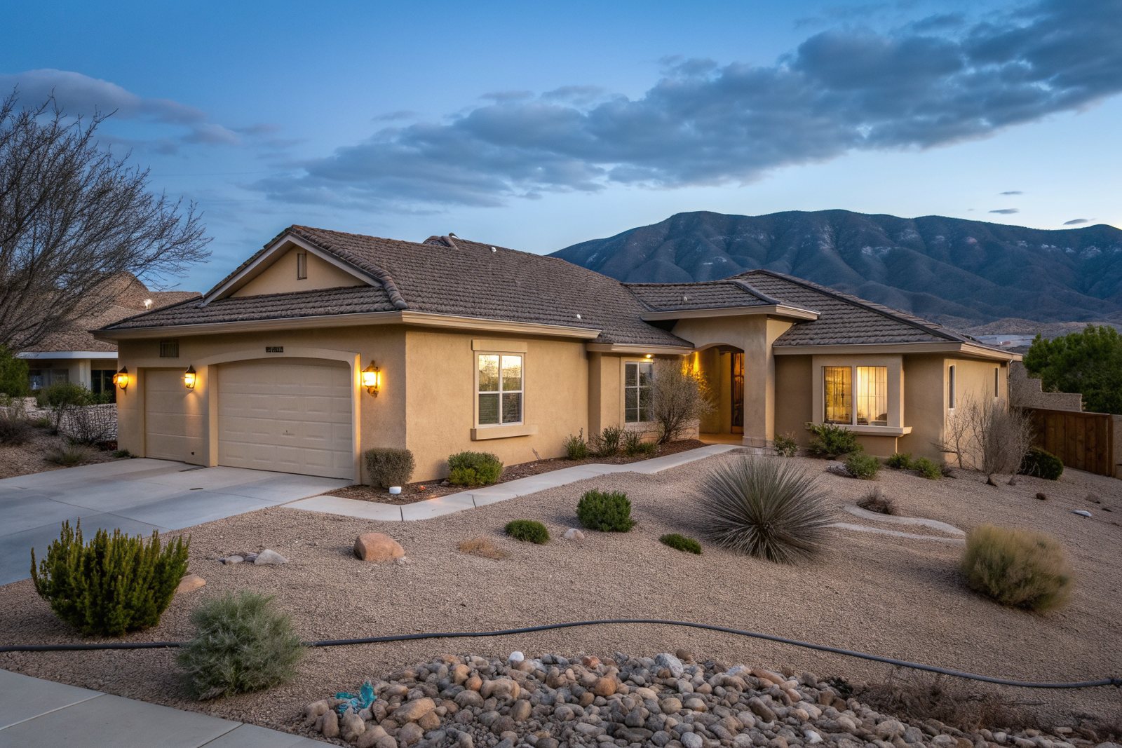 A tan stucco ranch home with a brown shingle roof and a gravel yard below the Sandia Mountains